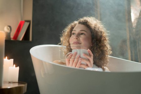 Téléchargez les photos : Femme aux cheveux longs souriante prend un bain avant d'aller au lit. Une femme agréable aux cheveux bouclés s'assoit dans un bain chaud entouré de bougies aromatiques et boit du thé chaud. Photo de haute qualité - en image libre de droit