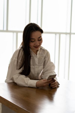 Foto de Una joven morena se sienta en una mesa en una habitación luminosa con un teléfono en las manos. Una morena con una camisa blanca está charlando con sus amigos vía messenger. Foto de alta calidad - Imagen libre de derechos
