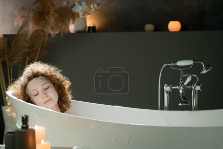 Photo for A beautiful woman with curly hair lies in a warm bath with a moisturizing mask on her face. A young woman takes a bath with essential oils. High quality photo - Royalty Free Image