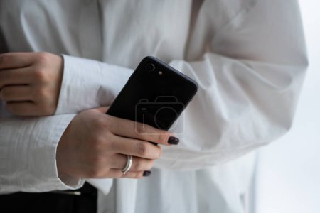 Photo for Cropped view of woman in white shirt holding smartphone in hand. A woman with a ring on her ring finger holds a black smartphone in her hand. High quality photo - Royalty Free Image