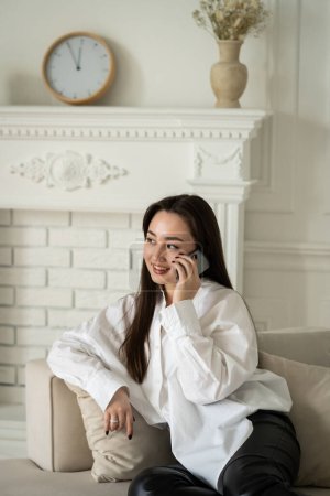 Photo for View of a young smiling woman who is sitting on the sofa and talking on the phone. A beautiful brunette discusses the details of the upcoming walk with her friend on the phone. High quality photo - Royalty Free Image