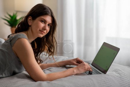 Photo for Smiling girl lying on the bed and using laptop with chroma key. Freelancer woman working in bedroom. High quality photo - Royalty Free Image