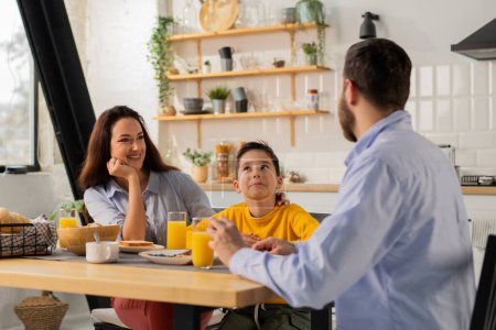 Téléchargez les photos : Un homme communique avec son fils et sa femme pendant le petit déjeuner. La famille est assise à la table de la cuisine avec des assiettes de sandwichs et des verres de jus d'orange. Photo de haute qualité - en image libre de droit