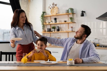 Photo for A smiling woman puts breakfast plates in front of her husband and son. A friendly family is going to have breakfast at the kitchen table. High quality photo - Royalty Free Image