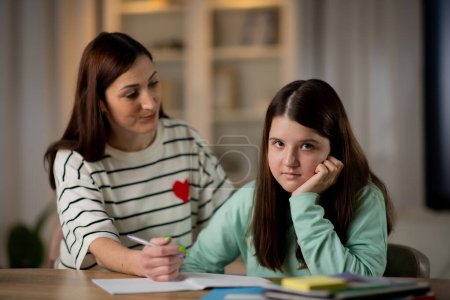 Téléchargez les photos : L'écolière ennuyée fait ses devoirs avec sa mère. Une jeune femme enseigne à sa fille à la maison. Éducation à domicile. Photo de haute qualité - en image libre de droit
