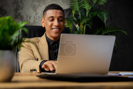 Téléchargez les photos : Homme d'affaires afro-américain souriant travaillant avec un ordinateur portable dans un bureau confortable avec des plantes. Avocat noir utilisant son ordinateur au bureau. Heureux entrepreneur dans un bureau moderne. Photo de haute qualité - en image libre de droit