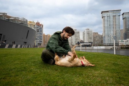 Foto de Un hombre barbudo, sentado en la hierba contra el telón de fondo de la ciudad, rasca el estómago de sus perros. Un hombre con una chaqueta verde juega con su perro mientras pasea. Foto de alta calidad - Imagen libre de derechos