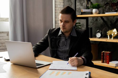 Photo for A young man in a business suit is working on a laptop while sitting in the office. A man in a black jacket sits in front of a laptop and fills out paperwork. High quality photo - Royalty Free Image