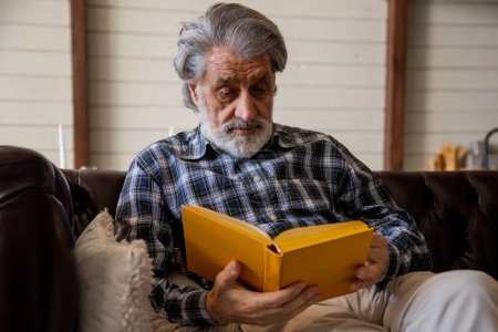 Téléchargez les photos : Homme âgé en chemise à carreaux livre de lecture tout en refroidissant dans le salon dans sa maison. Photo de haute qualité - en image libre de droit