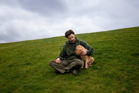 Foto de Un hombre se sienta con su perro en un césped verde. Un hombre barbudo abraza a un cocker spaniel mientras está sentado en la hierba contra un cielo nublado. Foto de alta calidad - Imagen libre de derechos