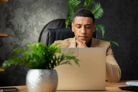Foto de Joven hombre afroamericano sosteniendo su barbilla con la mano y mirando la pantalla del portátil. Un hombre de negocios con un traje formal se sienta frente a una computadora en la oficina y charla con los negocios - Imagen libre de derechos