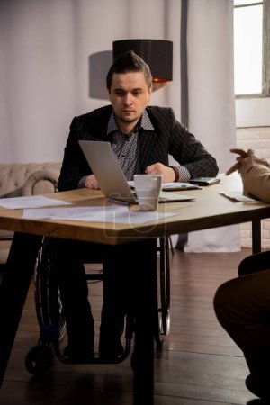 Foto de Un oficinista en silla de ruedas se sienta en una mesa con su colega y mira la pantalla de un portátil. La gente en trajes de negocios están sentados en una mesa con un ordenador portátil y varios documentos. Alta calidad - Imagen libre de derechos