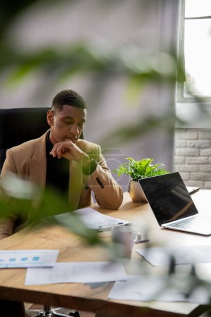 Téléchargez les photos : Un entrepreneur noir de sexe masculin est assis dans un bureau à domicile et étudie les états financiers de son entreprise. Un homme en costume officiel s'assoit à une table et étudie une pile de documents. Photo de haute qualité - en image libre de droit