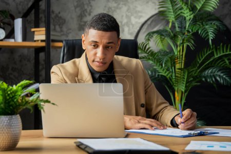 Téléchargez les photos : Un jeune homme noir est assis dans le bureau et regarde attentivement l'écran de l'ordinateur portable. Homme d'affaires assis au bureau à domicile signer des documents. Photo de haute qualité - en image libre de droit