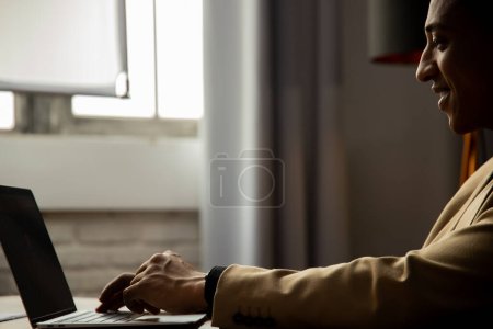 Téléchargez les photos : Vue latérale d'un homme vêtu d'une veste brune qui tape sur un clavier d'ordinateur portable. Homme d'affaires afro-américain souriant travaillant à distance à l'ordinateur à partir du bureau à domicile. Photo de haute qualité - en image libre de droit
