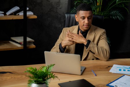 Foto de Un empresario afroamericano está sentado en un escritorio de su oficina. Un hombre con una chaqueta marrón se sienta frente a un portátil y piensa en algo. Foto de alta calidad - Imagen libre de derechos