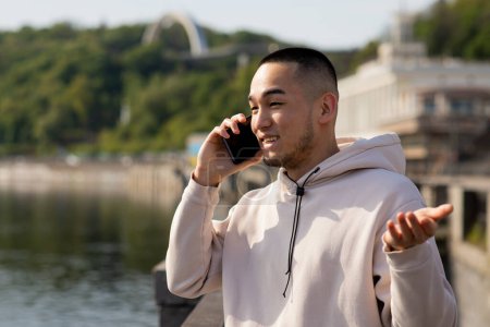 Photo for Young Asian man in sport hoodie is talking on the phone and smiling on the blurred nature background. High quality photo - Royalty Free Image