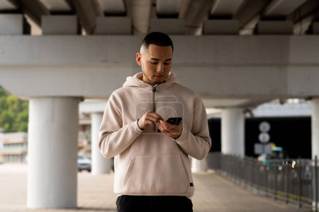 Foto de Un joven con rastrojo se destaca al aire libre y mira la pantalla de un teléfono inteligente. Hombre asiático está viendo algo en su teléfono mientras camina. Foto de alta calidad - Imagen libre de derechos