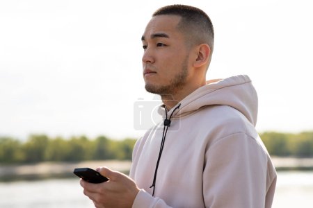 Photo for A young guy with a phone in his hands poses against a blurred background of nature. An Asian man stands in front of the camera holding a smartphone in his hand and looks away. High quality photo - Royalty Free Image