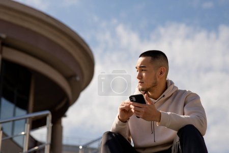 Téléchargez les photos : Un jeune Asiatique est assis à l'extérieur avec un téléphone dans les mains. Un homme s'assoit contre un ciel nuageux et regarde ailleurs. Photo de haute qualité - en image libre de droit