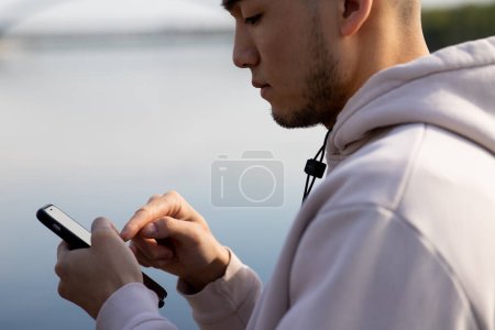 Téléchargez les photos : Un Asiatique se tient sur le fond de la rivière et tape sur l'écran du smartphone. Un jeune homme en sweat à capuche est debout à l'extérieur et tapant un message sur un smartphone. Photo de haute qualité - en image libre de droit