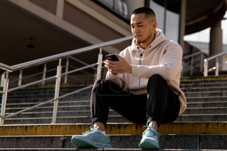 Téléchargez les photos : Un jeune Asiatique est assis sur les marches et tapant un message sur un smartphone. Un jeune homme utilise un smartphone assis à l'extérieur. Photo de haute qualité - en image libre de droit