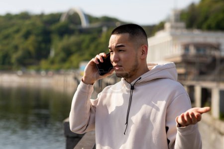 Photo for Sporty Asian man in hoodie is standing on the blurred background and talking on the phone. High quality photo - Royalty Free Image
