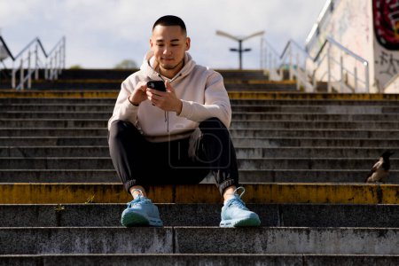 Photo for A man in a white hoodie sits on the steps and looks at something on his phone. Asian man sits outdoors and holds a telephone in his hands. High quality photo - Royalty Free Image