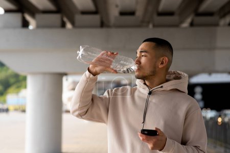Foto de Hombre asiático cansado con teléfono inteligente en sus manos es beber agua después de entrenar al aire libre. Foto de alta calidad - Imagen libre de derechos