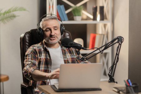 Foto de Hombre de pelo gris con camisa a cuadros está haciendo podcast. Teletrabajador está transmitiendo desde su habitación. Foto de alta calidad - Imagen libre de derechos
