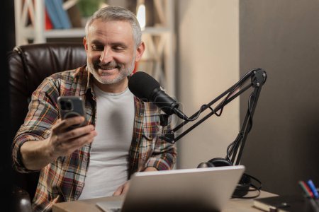 Photo for A gray-haired man with a beard is broadcasting on a streaming service. A male blogger broadcasts live and reads questions from viewers from his phone. High quality photo - Royalty Free Image