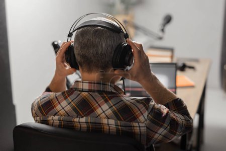 Photo for A man in a plaid shirt sits at a table in front of a laptop and a microphone. A man with gray hair puts on headphones and prepares to record a new podcast. High quality photo - Royalty Free Image
