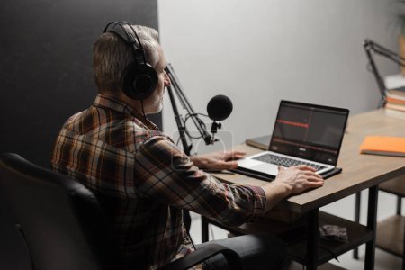 Photo for A man in headphones records a podcast using a studio microphone and a laptop. A gray-haired man in a plaid shirt sits at a table and looks at a computer screen. High quality photo - Royalty Free Image