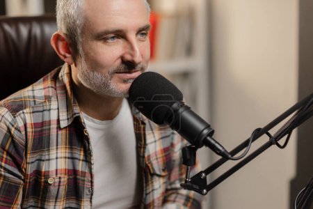 Photo for A bearded man in a plaid shirt sits at a table in front of a studio microphone. Male blogger recording a podcast using a professional microphone. High quality photo - Royalty Free Image