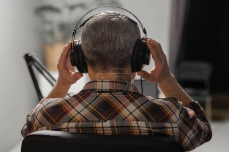 Photo for Rear view of a gray-haired man who sits in a chair and adjusts the headphones on his head. A man in a plaid shirt sits in his room and prepares to record a podcast. High quality photo - Royalty Free Image