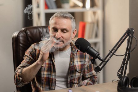 Téléchargez les photos : Un homme fume une cigarette dans un studio de radio. Un blogueur masculin s'assoit à une table devant un microphone de studio et fume une cigarette. Photo de haute qualité - en image libre de droit