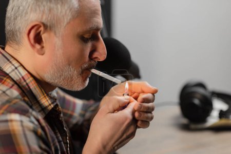Foto de Vista lateral de un hombre de pelo gris con un cigarrillo. Un hombre con una camisa a cuadros se sienta en una mesa y enciende un cigarrillo. Foto de alta calidad - Imagen libre de derechos