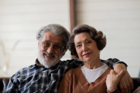 Téléchargez les photos : Belle famille âgée souriante couple mari et femme regardent la caméra avec tendresse et amour tout en étant assis dans le salon à la maison, retraité homme et femme étreignant à l'intérieur. Photo de haute qualité - en image libre de droit