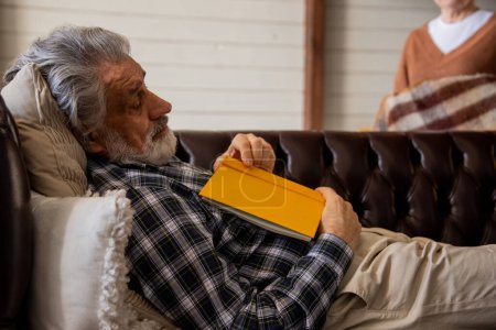 Téléchargez les photos : Vieux lecteur masculin faisant la sieste à canapé confortable à la maison.Grand-père s'est endormi sur le canapé tout en lisant. Photo de haute qualité - en image libre de droit