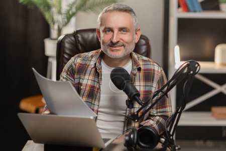 Foto de Un blogger sonriente posando para la cámara mientras está sentado en su lugar de trabajo. Un hombre de pelo gris con barba se sienta en una mesa frente a un portátil y un micrófono y mira a la cámara. Alta calidad - Imagen libre de derechos