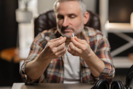 Photo for A bearded man with gray hair sits at a table and holds a broken cigarette in his hands. High quality photo - Royalty Free Image