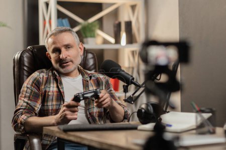 Photo for A gray-haired man in a plaid shirt sits in front of the camera and holds a gamepad in his hands. A male tech blogger is recording a video review of a new gamepad. High quality photo - Royalty Free Image