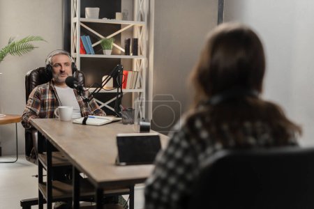 Photo for A man in headphones sits at a table opposite a woman and says something into a microphone. Two bloggers are recording a joint podcast from the studio. High quality photo - Royalty Free Image