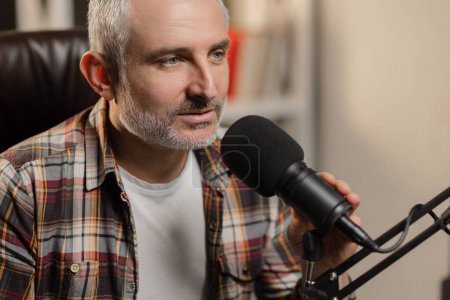 Foto de Un hombre con el pelo gris se sienta en una mesa y ajusta un micrófono de estudio. Un blogger masculino está grabando un podcast usando un micrófono profesional. Foto de alta calidad - Imagen libre de derechos