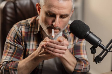 Photo for A bearded man sits at a table in front of a microphone and lights a cigarette. A gray-haired man in a plaid shirt smokes while sitting at the table. High quality photo - Royalty Free Image