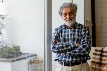 Photo for A gray-haired man in a shirt stands against the background of a large window. A smiling, gray-haired man with a beard stands idly in the house. High quality photo - Royalty Free Image