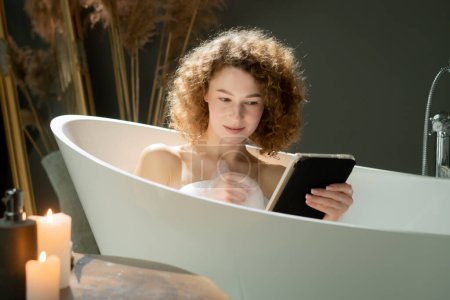 Foto de Jovencita atractiva está tumbada en el baño y viendo algo en una tableta digital. Una mujer hermosa está navegando en las redes sociales en su tableta mientras toma un baño nocturno. Foto de alta calidad - Imagen libre de derechos