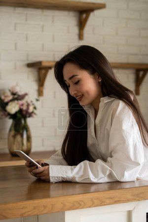 Photo for A beautiful brunette sits at a table and smiles while watching funny videos on her phone. Smiling woman in a white shirt is using a smartphone. High quality photo - Royalty Free Image