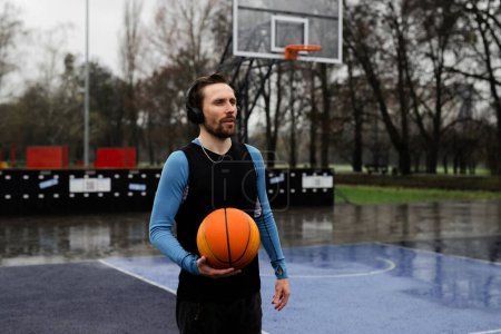 Photo for Concept of rainy weather crossfit exercises. Sportsman prepare to throw ball in hoop. Basketball player man warmup before training at sport playground. Athletic guy listen wireless headphones on local - Royalty Free Image