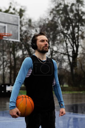 Photo for Strong amateur sportsman playing basketball when raining weather. Young guy listen music wireless headphones for workouts. Man in sportswear using earphones on outdoor playground background. Hobby - Royalty Free Image
